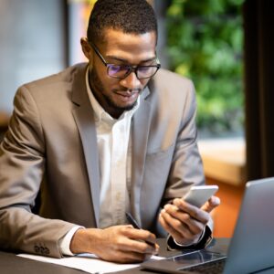 Black business guy taking notes in notebook holding phone